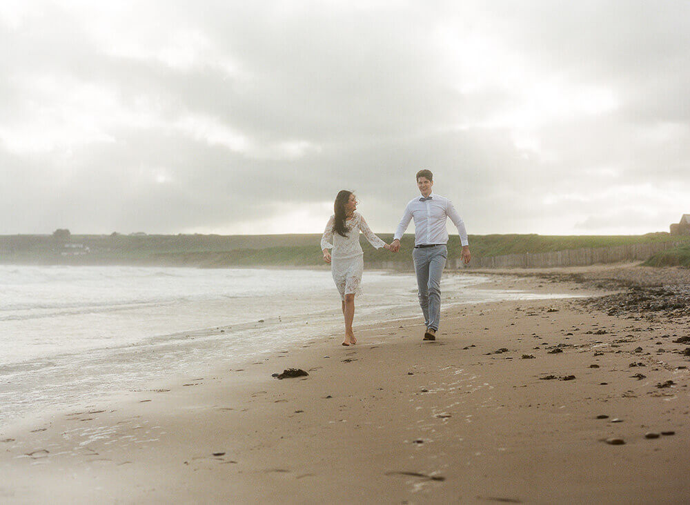 A bride and groom holding hands and running on a sandy misty beach in Ireland. Photo by AMBphoto