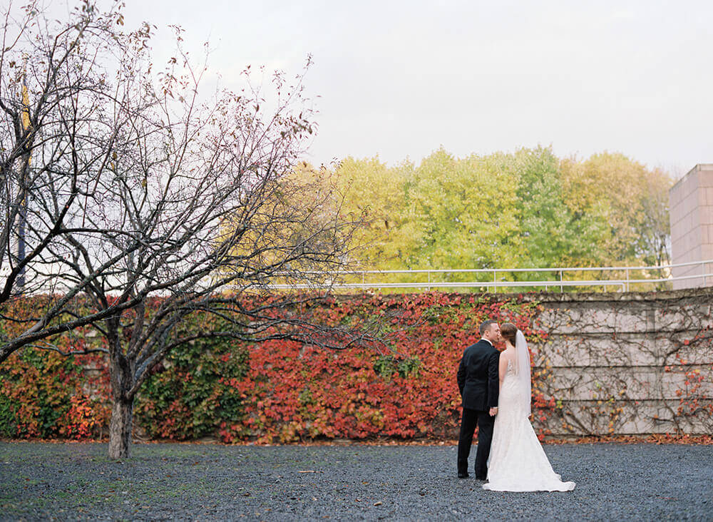 A bride and groom standing in front of a downtown Ottawa stone wall covered in fall leaves and colours on their wedding day. Photo by AMBphoto