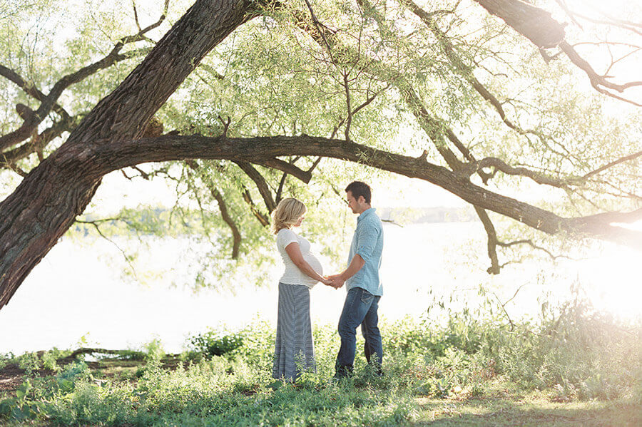 A pregnant woman and her husband stand togehter beneath a tree on the banks of the Ottawa river as the sun breaks through the branches. Photo by AMBphoto